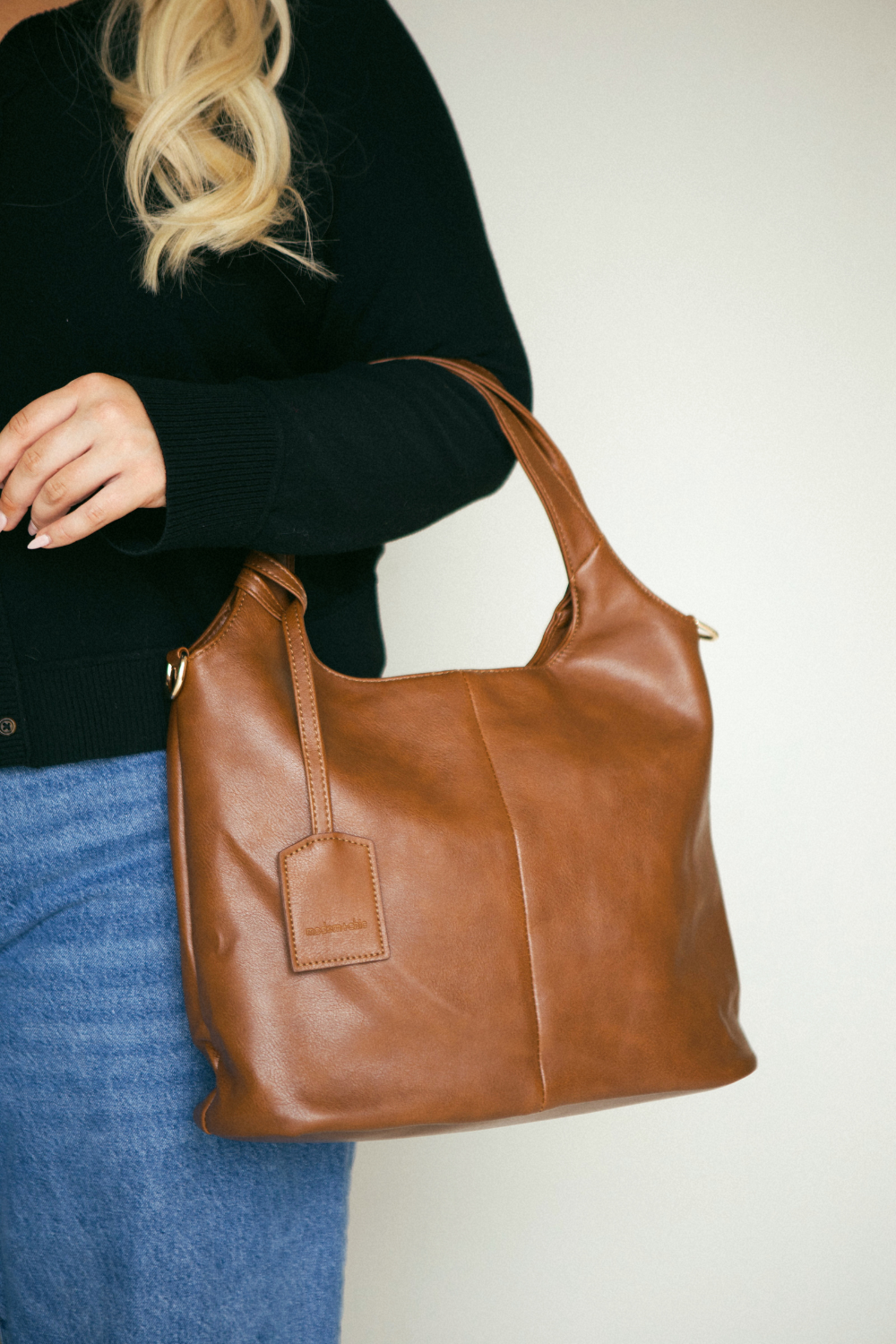 Woman wearing jeans and a patterned jacket holding a camel Modern+Chic vegan leather Clara slouchy tote in front of her body hanging down to her knees. 