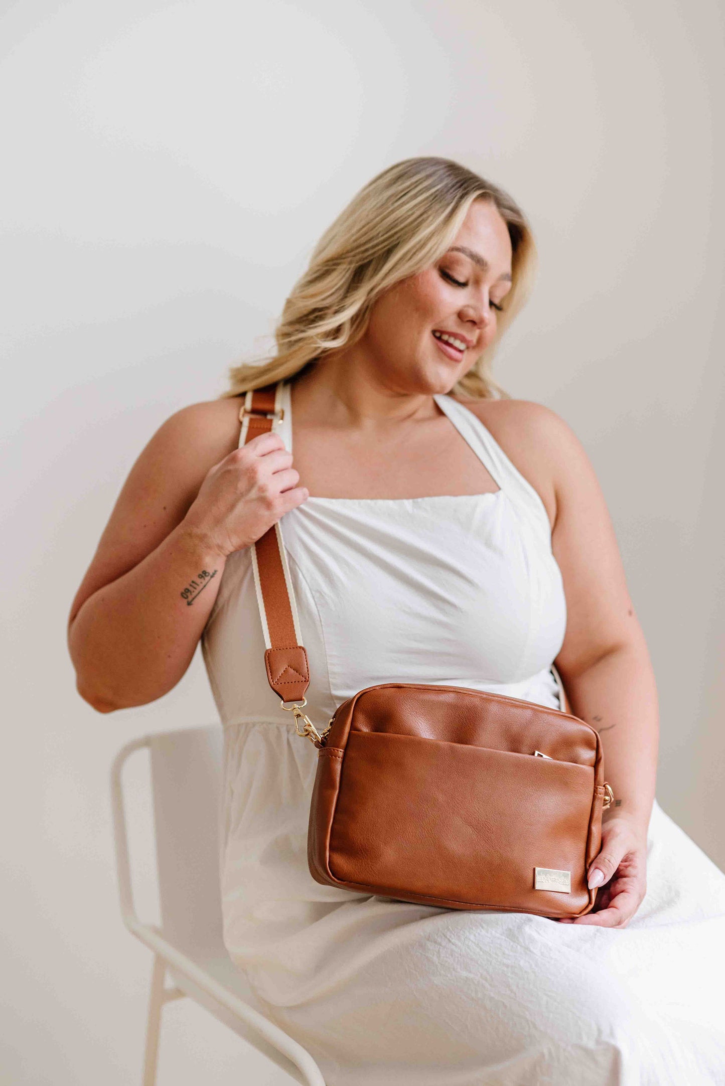 A blond haired woman modeling a camel colored Modern + Chic Alice Crossbody Vegan Leather bag. 