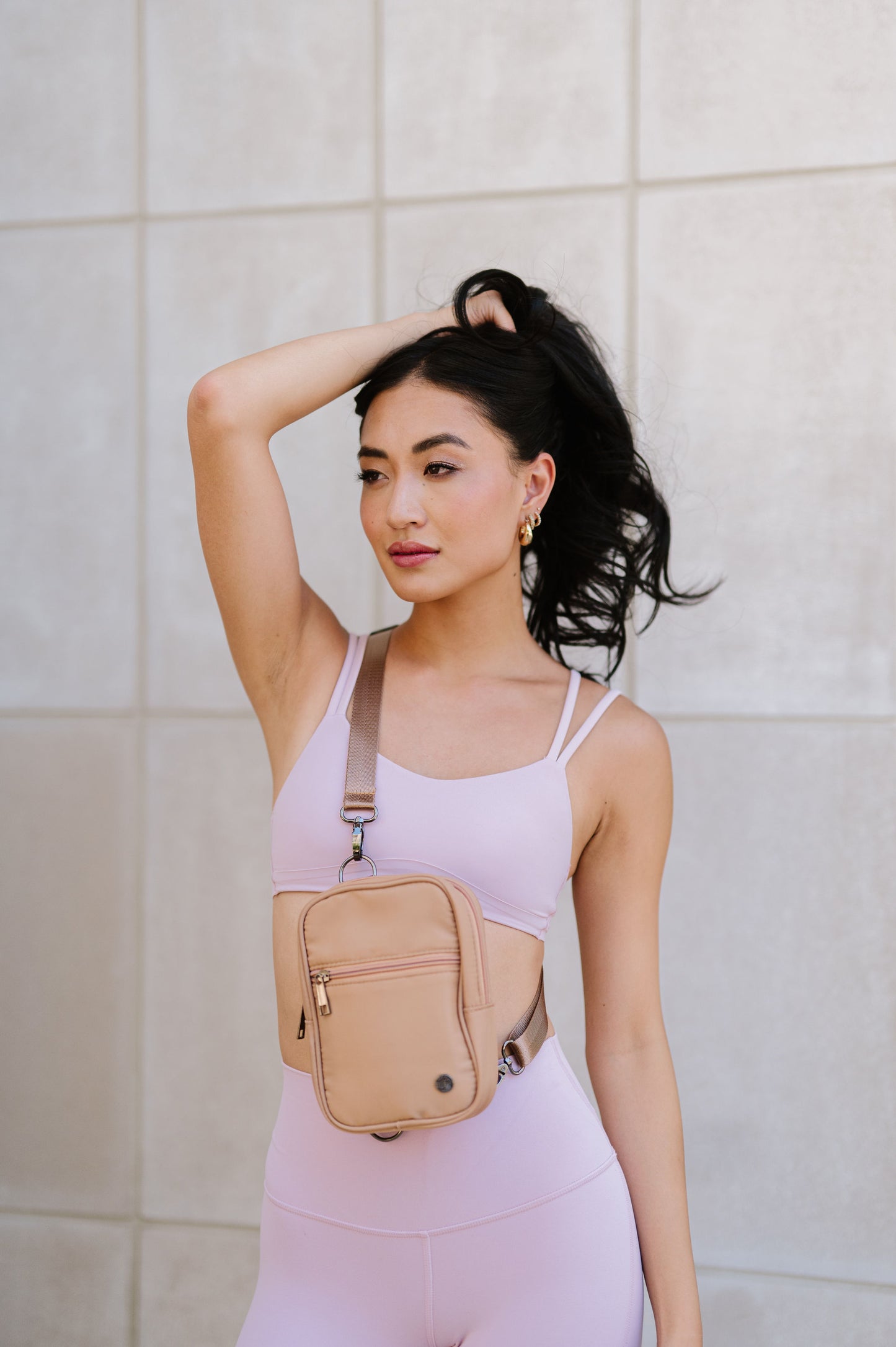 A woman wearing a pink sports bra and leggings showing a Modern + Chic Lainey Convertible Crossbody Bag for Women on crossbody. 