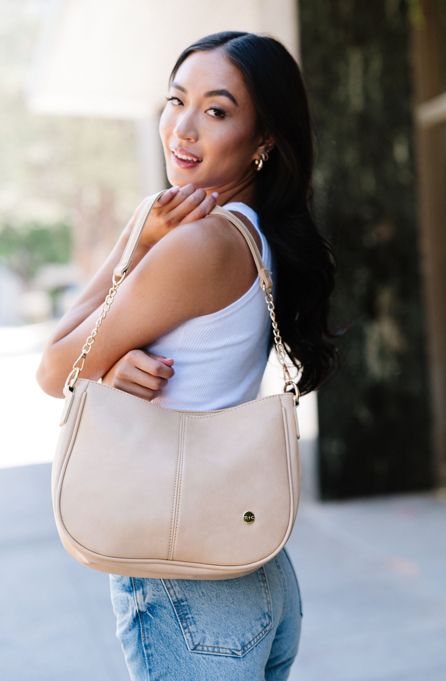 A woman wearing a cream Modern + Chic Marissa Vegan Leather Crossbody handbag over her shoulder displaying the gold chain accents on the strap.
