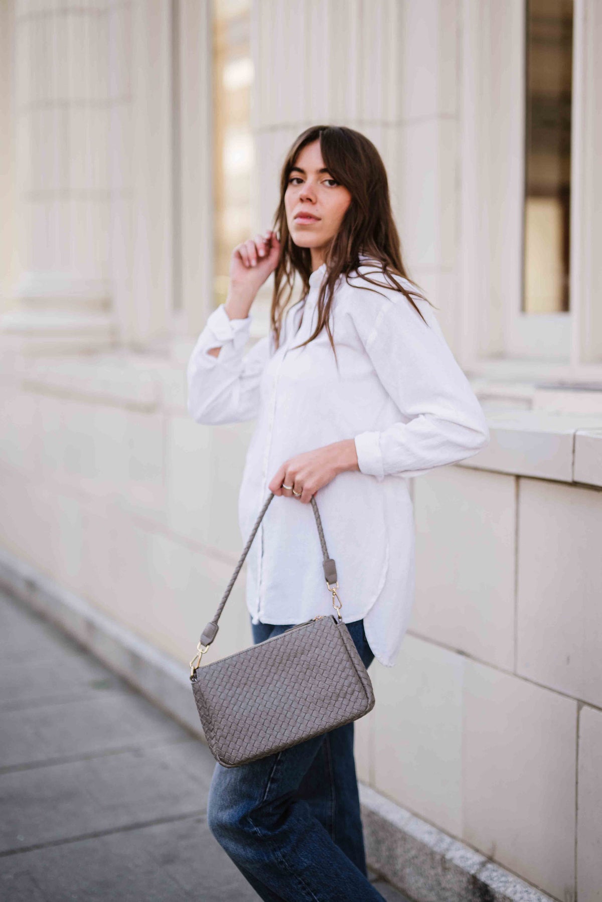 Woman in white shirt and jeans holding a gray Modern + Chic Elle Woven Shoulder Bag for Women against a light-colored building.