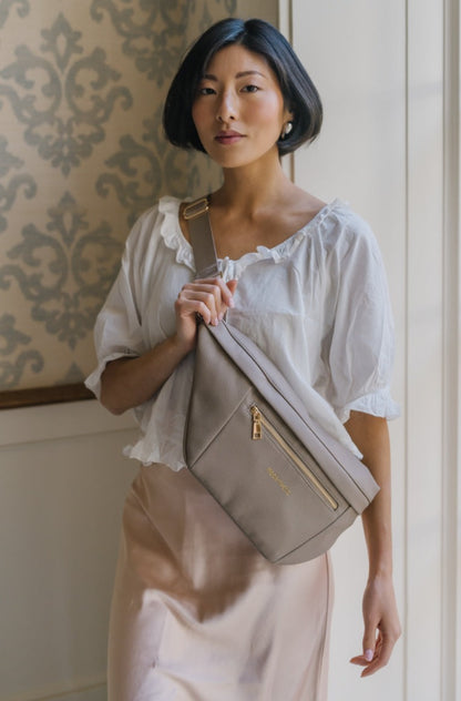 A woman wearing an oversized taupe Modern+Chic Presley Oversized Sling Bag for Women with gold hardware across her torso.