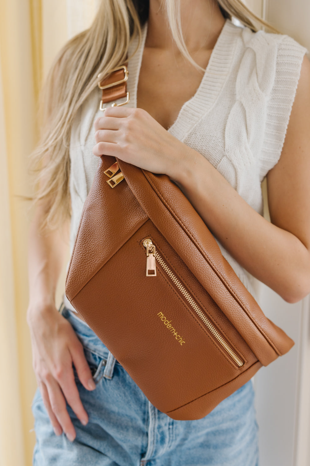 A woman wearing a white sweater vest and an oversized camel Modern+Chic Presley Oversized Sling Bag for Women with gold hardware across her torso.