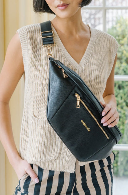 A woman wearing a cream sweater vest and an oversized black Modern+Chic Presley Oversized Sling Bag for Women with gold hardware across her torso.