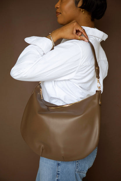 Person wearing a white shirt and blue jeans, holding a brown vegan leather Modern+Chic Savannah Slouchy Tote against a brown background. 