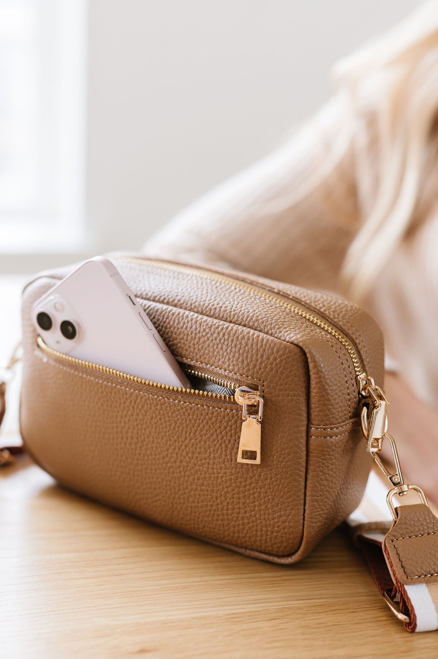 A beige Modern+Chic Willow Vegan Leather Convertible Crossbody Bag sitting on a table with a pink phone partially showing out of the unzipped back pocket. 