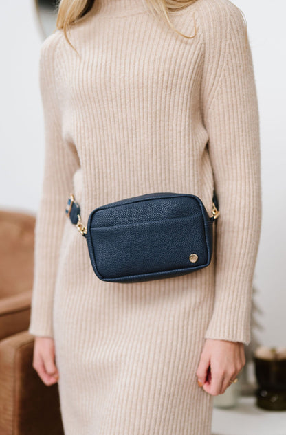 Woman wearing a blue Modern+Chic Willow Vegan Leather Convertible Crossbody Bag around her waist as a belt bag on a white background. 